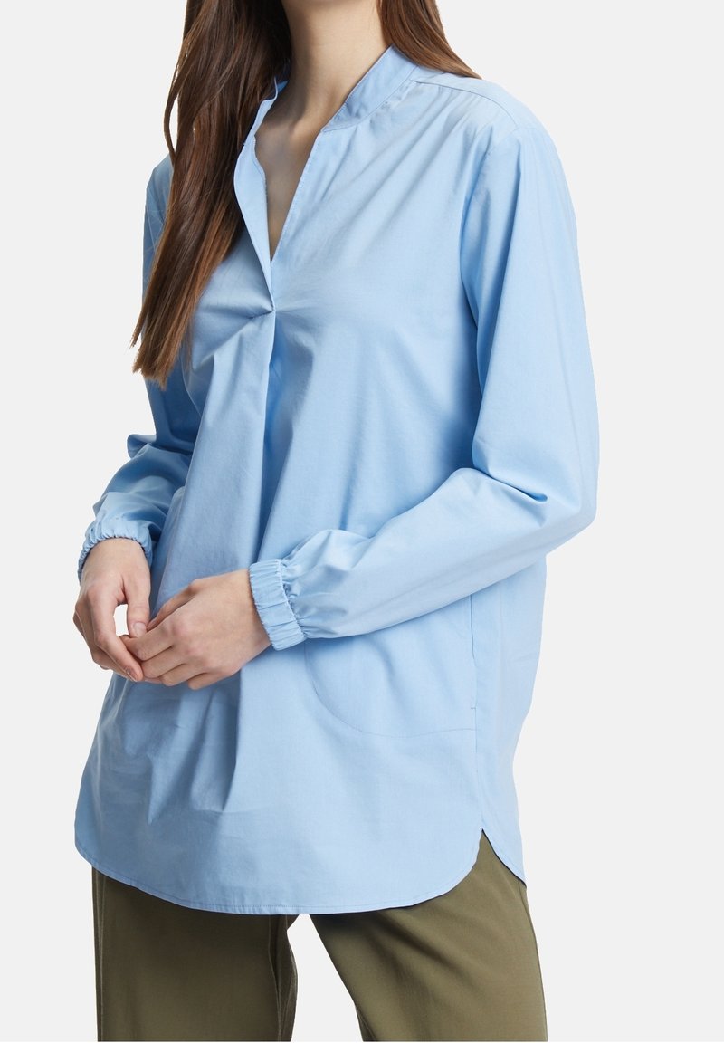 Jasnoniebieska tunika z bawełny, z stójką, długimi rękawami, elastycznymi mankietami, bocznymi kieszeniami i zaokrąglonym dołem.