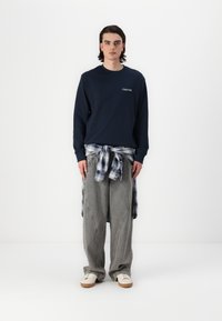 Marineblauer Sweatshirt mit "Club1990"-Logo, graue gestreifte weite Hose und karierte Hemd um die Taille gebunden; weiße Schuhe mit Gummisohlen.