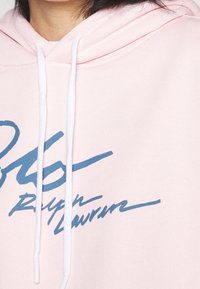 Ljusrosa hoodie med vita dragsnören och en blå kursiv logotyp som står "Ralph Lauren." Mjukt tyg med en slät textur.