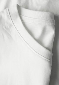 Zložena bela bombažna majica z vidnim rebrastim okroglim ovratnikom in prišitimi šivi na enobarvnem ozadju.