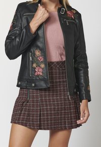 Veste de motard en cuir noir avec broderies florales en rouge et vert, poches zippées et design ajusté. Associée à une jupe à carreaux.