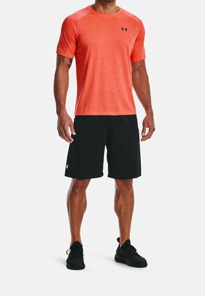 Uomo che indossa una maglietta sportiva a maniche corte di colore arancione brillante, pantaloncini sportivi neri e scarpe da corsa nere, in piedi con le braccia rilassate.