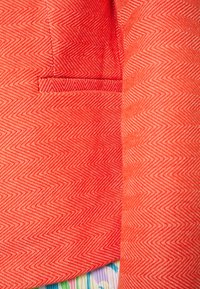 Rood visgraatpatroon stof met een klein zakdetail, met een gestructureerde afwerking en een soepele vallende stof. Een kleurrijke stof piept eronderuit.