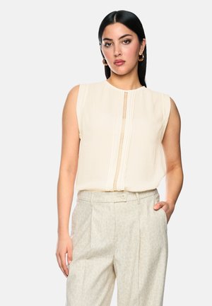 Woman with straight black hair wears sleeveless beige blouse with lace detail and light patterned high-waist trousers, hand in pocket.