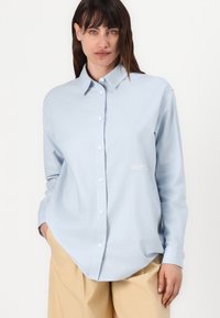 Lyseblå skjorte med knapper laget av bomull. Har klassisk krage, lange ermer og brodert merkevare på venstre bryst.