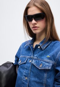 Νεαρό άτομο που φοράει μαύρα γυαλιά ηλίου τύπου wraparound και μπλε τζιν μπουφάν με μικρό λογότυπο στο στήθος, κρατώντας ένα μαύρο δερμάτινο αντικείμενο.