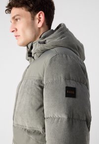 Grå puffer jakke med ribbet tekstur, hætte og et sort emblem med "BOSS" logo på ærmet for ekstra detaljer.
