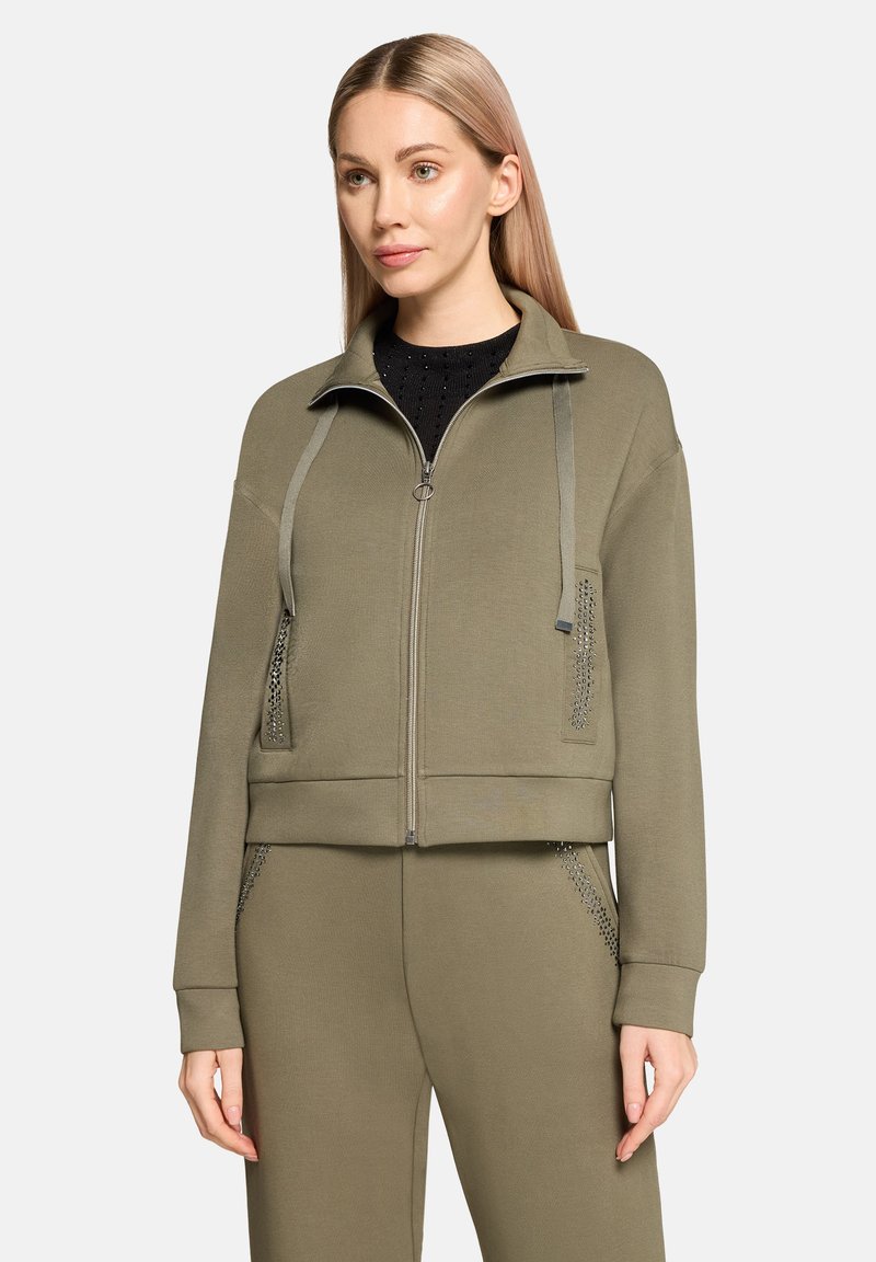 Krótka kurtka khaki zapinana na zamek z teksturowanymi akcentami i detalem ze sznurkiem. Miękki materiał, kieszenie boczne ozdobione ćwiekami.