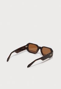 Lunettes de soleil écaille de tortue avec une forme octogonale, des verres marron et des détails dorés sur les branches. Monture en acétate texturée avec une finition brillante.