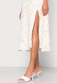 Vestido midi floral con base blanca que presenta flores en tonos pastel, abertura lateral y combinado con sandalias deslizantes blancas. Textura suave y tela liviana.