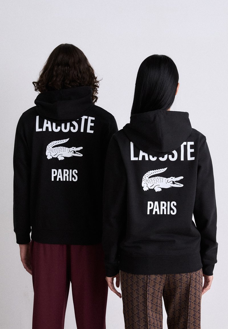 Juodi kapučiniai su baltais užrašais "LACOSTE" ir "PARIS", su krokodilo grafiniais vaizdais ant nugaros. Minkšta medžiaga, laisvas kirpimas ir standartinės rankovės.