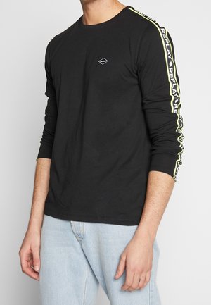 Mand iført sort langærmet skjorte med "Replay"-logoer på brystet og gule kanter på ærmerne, kombineret med lyseblå jeans, mod en hvid baggrund.