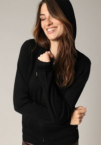 Zwarte hoodie gemaakt van zacht materiaal, met een rits aan de voorkant, lange mouwen en geribbelde manchetten. Subtiele glanzende accenten door het materiaal.