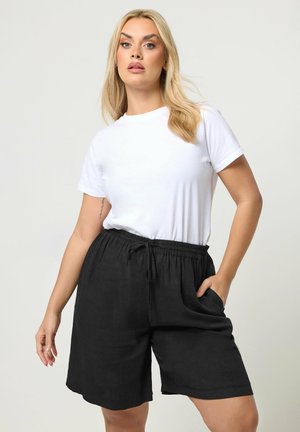 Blonde Frau mit offenem Haar, die ein weißes kurzärmeliges Hemd und schwarze Shorts mit Kordelzug trägt, posiert mit einer Hand in der Tasche.