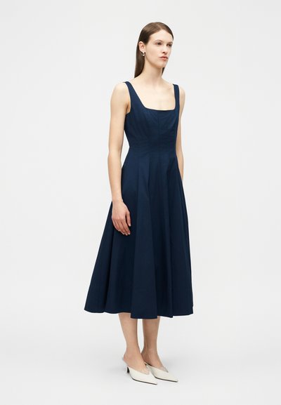 Γυναίκα που φοράει ένα μπλε ναυτικό μίντι φόρεμα χωρίς μανίκια με τετράγωνο ντεκολτέ και λευκά χαμηλά παπούτσια με μυτερή μύτη, στέκεται μπροστά σε απλό φόντο.