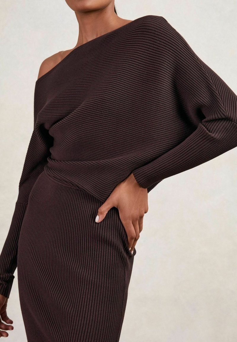 Femeie îmbrăcată cu pulover din tricot ribbed, cu un umăr descoperit, de culoare maro închis, și fustă asortată, cu mâna plasată pe șold, pe un fundal neutru.