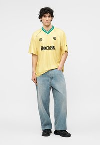 Mladić stoji, nosi žutu Barrow majicu sa zelenim obrubom na ovratniku, lepršave svijetloplave traperice i crne tenisice, neutralna pozadina.