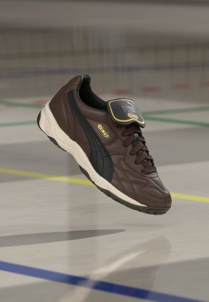 Καφέ και μαύρο αθλητικό παπούτσι Puma με λευκή σόλα που αιωρείται πάνω από ένα γυμναστήριο με πολύχρωμες γραμμές στο δάπεδο.