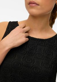 Czarna koronkowa sukienka z teksturowanym wzorem, okrągłym dekoltem i bezrękawowym krojem. Zbliżenie na rękę na obszarze ramienia.