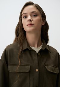 Giacca camicia in suede marrone con due tasche frontali, chiusura con bottoni e bottoni in tonalità dorata. Maniche lunghe e colletto.