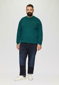 Groene textuurtrui met ronde hals, gecombineerd met donkerdenim jeans en witte sneakers. Bevat een logo aan de linkerkant van de borst.