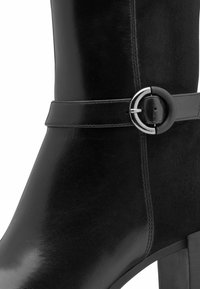 Bota de cuero negro hasta el tobillo con un acabado suave, puntera redondeada y una correa decorativa con hebilla. El tacón es alto y de forma cuadrada.