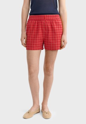 Osoba v červených károvaných šortkách s elastickým pásom a dvoma gombíkmi, béžové baleríny a tmavomodrý bezrukávový top.