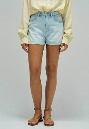 Personne portant un short en denim bleu clair, une chemise à manches longues boutonnée jaune pâle et des sandales plates marron à lanières, debout devant un fond uni.