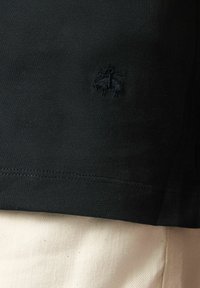 Černé krátké tričko s krátkým rukávem z elastického materiálu, s malým vyšitým logem v tmavěmodré barvě na levé straně hrudi. Viditelné švy.