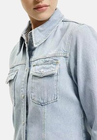 Jasnoniebieska kurtka dżinsowa z kołnierzykiem, widocznymi szwami, dwoma przednimi kieszeniami i zapięciem na guziki. Materiał ma klasyczną teksturę dżinsu.