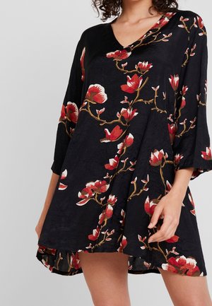 Robe noire ample avec imprimé floral rouge et blanc, col en V et manches trois-quarts, portée par une personne aux cheveux bouclés.