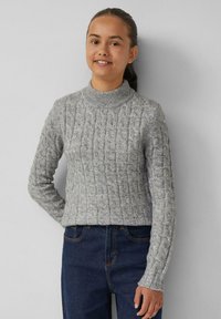 Grauer Strickpullover mit hohem Kragen, versehen mit einem strukturierten Zopfmuster. Kombiniert mit blauen Jeans.