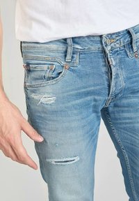 Lichtblauwe denim jeans met een verouderd ontwerp en zichtbare scheuren. Koperkleurige hardware en een slim fit vorm.