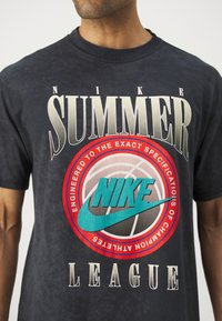 Marineblå bomull T-skjorte med dristig hvit og rød tekst: "NIKE SUMMER LEAGUE." Inkluderer stor tealfarget Nike-logo innenfor et sirkeldesign.