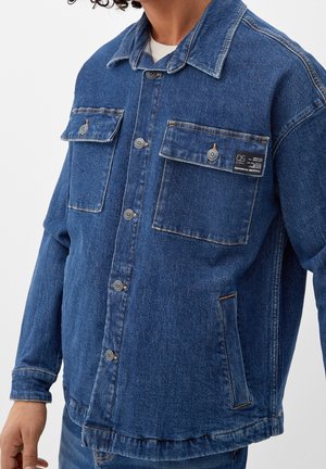 Blå denimjakke med to brystlommer, kontrastknapper og et mærke på venstre lomme. Har en knappet front og klassisk krave.