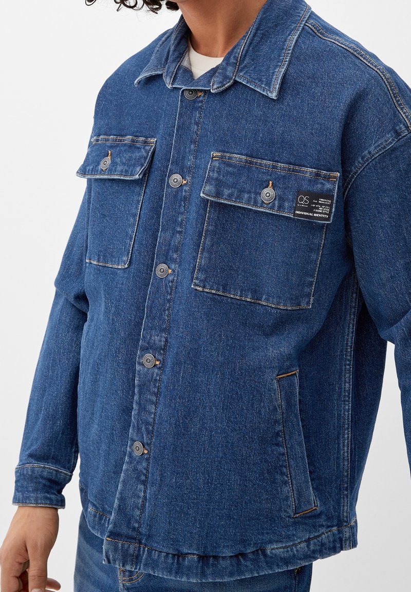 Chaqueta de mezclilla azul con dos bolsillos en el pecho, botones en contraste y una etiqueta en el bolsillo izquierdo. Presenta un cierre delantero con botones y un cuello clásico.