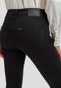 Zbliżenie osoby noszącej czarne jeansy Lee i białą koszulę z długim rękawem oraz mankietami na guziki, ukazujące szczegóły tylnej kieszeni.