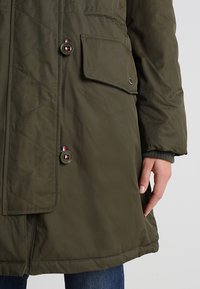 Manteau matelassé vert olive avec poche à rabat et boutons décoratifs, porté avec un jean bleu, main reposant sur le côté.