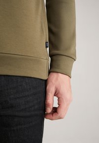 Felpa verde oliva con polsini a costine, spacco laterale e etichetta del logo, abbinata a jeans scuri, evidenziando la texture del tessuto e il contrasto dei colori.