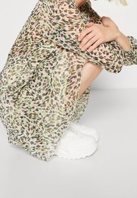 Průhledné šaty se vzory v světlé zelené, růžové a hnědé, s volným střihem a nařasením, nošené s bílými masivními teniskami.