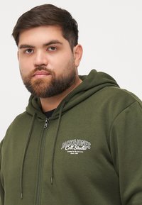 Maslinasto zeleni zip-up dukserica od mekog materijala, s bijelim logom na lijevoj sisi, sa vezicama i teksturiranim završetkom.
