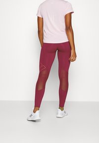 Bordeaux leggings met mesh-inzetten, hoge taille, gladde textuur, gecombineerd met een lichtroze top met korte mouwen. Witte sportschoenen.