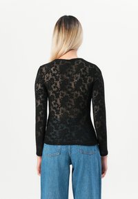 Kvinne med blondt hår har på seg en langermet svart blomstret blonder topp og blå høytaljede jeans, stående vendt bort fra kameraet mot en hvit bakgrunn.