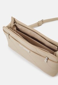 Beige křížová taška s prošívaným designem, stříbrnými detaily, zipovými přihrádkami a texturovaným povrchem. Součástí je nastavitelné rameno.
