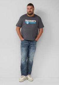 Graues Baumwoll-T-Shirt mit dem Logo des "Discovery Channel" in Weiß und Blau, kombiniert mit hellblauen Jeans und beigen Sneakers. Stehende Pose.
