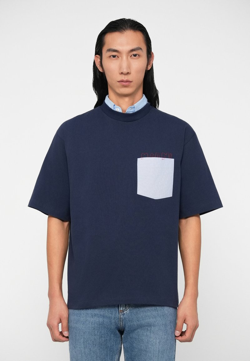 Marineblaue Baumwoll-T-Shirt mit kurzen Ärmeln, ausgestattet mit einer hellblauen Tasche und dezentem roten Text-Detail auf der Brust. Lässige Passform, minimalistisches Design.