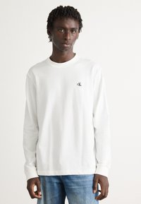 Ung mann med korte dreads iført en hvit longsleeve-skjorte med en liten "cK" logo og blå jeans, stående foran en ensfarget bakgrunn.