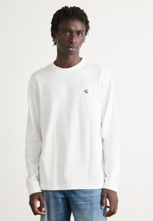 Ung mand med korte dreads iført en hvid langærmet skjorte med et lille "cK" logo og blå jeans, stående foran en ensfarvet baggrund.