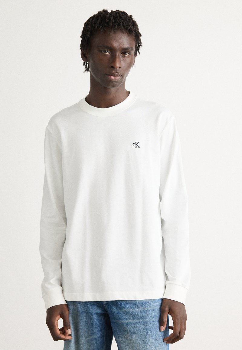 Ung mann med korte dreads iført en hvit longsleeve-skjorte med en liten "cK" logo og blå jeans, stående foran en ensfarget bakgrunn.