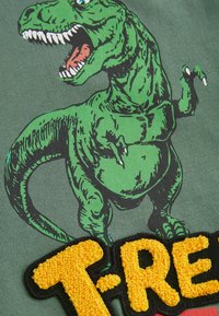 Grünes T-Rex-Graphic auf einem grauen Shirt mit schwarzen Konturen; strukturierter, gelber, gestickter "T-REX"-Patch deutlich sichtbar.
